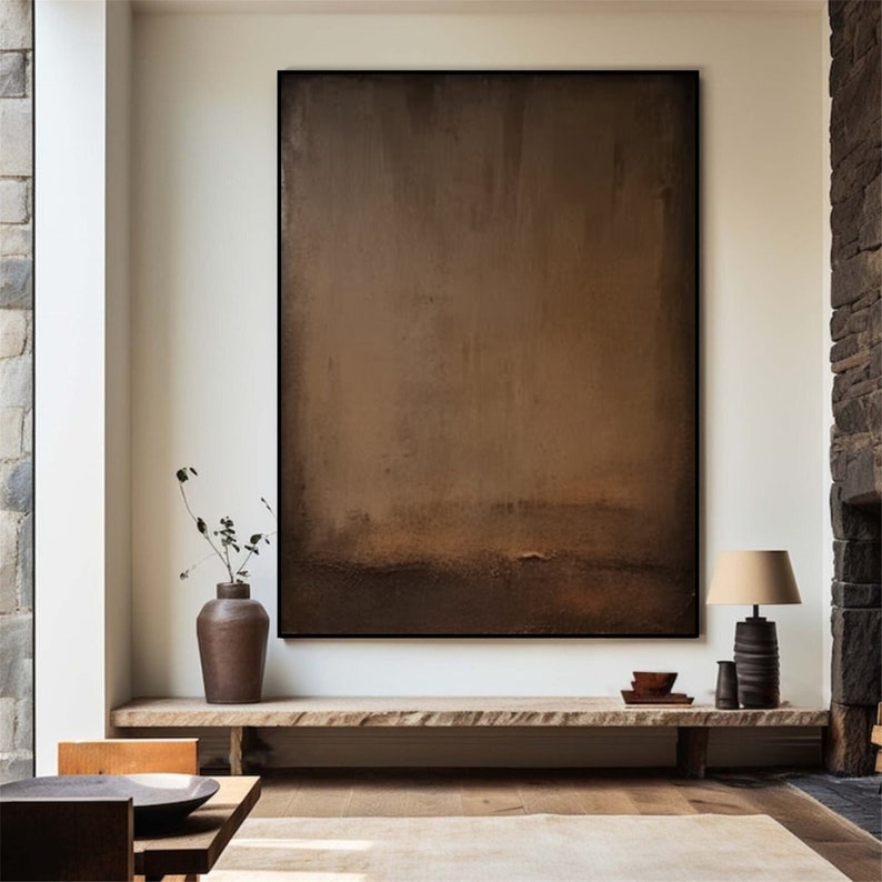 May include: A large, framed abstract painting in shades of brown and tan dominates the room. A wooden shelf displays a vase with greenery, a lamp, and decorative bowls. The room has a neutral color palette with a stone fireplace.