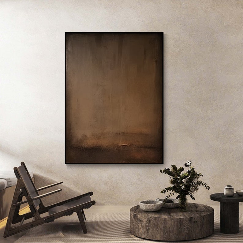 May include: Abstract brown painting in a black frame. The artwork features a blend of brown tones, creating a textured, minimalist aesthetic. The painting is displayed in a modern living room setting with a wooden chair and a round coffee table.