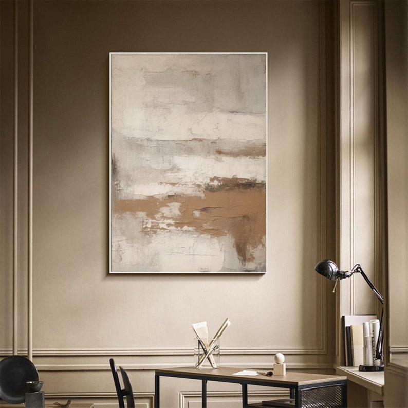May include: Abstract painting with brown and white brushstrokes on a canvas. The painting is framed in a white frame and is hanging on a beige wall.