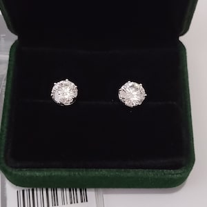 Puede incluir: Un par de pendientes de botón de plata con piedras redondas de circonita cúbica transparentes engastadas en garras. Los pendientes se muestran en una caja de joyería de terciopelo verde.