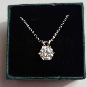 Könnte beinhalten: Eine silberne Halskette mit einem großen, runden, klaren Edelsteinanhänger. Der Anhänger ist in einer sechszackigen Fassung gefasst und hängt an einer zarten Kette. Die Halskette wird in einer dunkelgrünen Schmuckschatulle präsentiert.