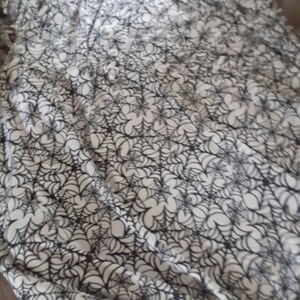 Puede incluir: Una manta blanca y negra con estampado de telara&ntilde;as. La manta presenta un dise&ntilde;o repetido de telara&ntilde;as negras sobre un fondo blanco. Los bordes de la manta est&aacute;n acabados con flecos anudados.
