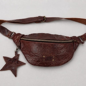 May include: A dark brown leather bum bag with a textured, crinkled surface, a single zippered main compartment, and an adjustable strap with a metal buckle. A matching leather star-shaped charm hangs from the zipper pull on the left side.