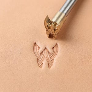 May include: A close-up of a leather stamping tool with a decorative design. The tool is made of brass and steel, and is pressed into a piece of tan leather, creating an embossed pattern. The embossed pattern is a stylized, symmetrical design.