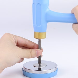 K&ouml;nnte beinhalten: Ein blauer Gummihammer mit Messingkopf wird verwendet, um einen Metallpunzen in eine Metallbasis mit einem blauen Gummiring zu treiben.