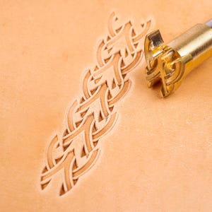 Könnte beinhalten: Ein Lederstempelwerkzeug aus Messing mit einem komplexen geometrischen Musterkopf, das neben einem passenden geprägten Design auf einem Stück hellbraunem, pflanzlich gegerbtem Leder liegt. Das Werkzeug hat einen zylindrischen Metallschaft.