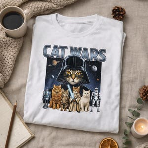 May include: White t-shirt with a "CAT WARS" graphic. The design features a cat in a Darth Vader helmet, surrounded by other cats dressed as Star Wars characters. A space scene is in the background.