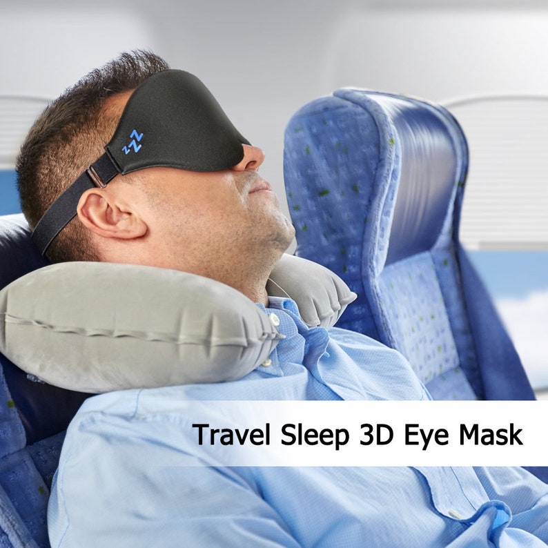 Op de afbeelding: Een man met een zwart slaapmasker met blauwe stiksels en een grijs reiskussen slaapt in een vliegtuigstoel. De tekst "Travel Sleep 3D Eye Mask" is zichtbaar onderaan de afbeelding.
