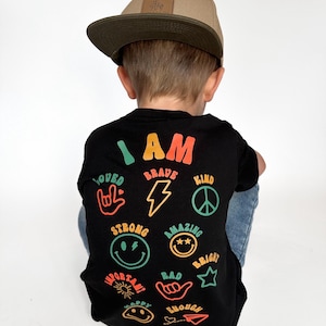 May include: A black t-shirt with the words "I AM" in colorful letters and various positive words and symbols printed on the back. The words include "loved", "brave", "kind", "strong", "amazing", "bright", "important", "rad", "happy", and "enough".