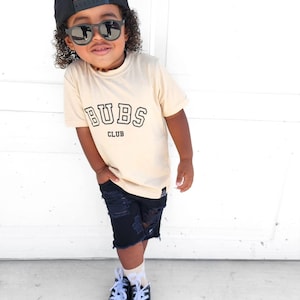 May include: A young child wearing a beige t-shirt with the text "BUBS CLUB" in black, black sunglasses, a black baseball cap, ripped denim shorts, and black and white sneakers.