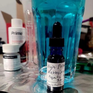 Może przedstawiać: Mała, ciemnoniebieska szklana butelka z czarną pipetą, z napisem "High Potency Methylene Aquatic 1m Handcrafted". Przezroczysty niebieski płyn znajduje się w szklance za butelką. Obok butelki leży moneta.