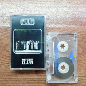 May include: A vintage cassette tape with a black and white album cover featuring the band Pulp. The album is titled "Different Class". The cassette is in a clear plastic case, and the tape is visible inside.