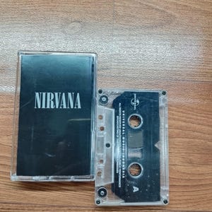 Op de afbeelding: Een doorzichtige cassetteband met een zwarte inleg met de bandnaam "NIRVANA" in het wit. De cassetteband is zichtbaar in de behuizing, met het label "UNIVERSAL MUSIC-INDONESIA". De behuizing is open en ligt op een houten oppervlak.