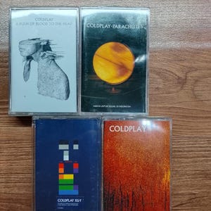 May include: Four vintage Coldplay cassette tapes. The album covers feature various designs, including a goat illustration, a globe, a colorful geometric design, and an orange textured background. Text on the tapes includes album titles and promotional notes.