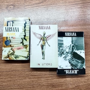 Op de afbeelding: Drie Nirvana album-cd's worden getoond. De albumhoezen zijn onder meer "Sliver", "In Utero" en "Bleach". De "In Utero" cd toont een naakte engelenfiguur met vleugels. De "Bleach" cd is zwart-wit.
