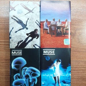 Puede incluir: Cuatro álbumes en CD de la banda Muse. Las portadas presentan varios diseños, incluyendo un hombre con sombras, un grupo de hombres en una mesa, hongos azules brillantes y una mujer con un vestido blanco. Los álbumes se titulan "Greatest Hits" y "Showbiz".