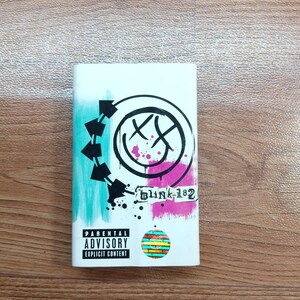 May include: A white rectangular item featuring the band Blink-182 logo with a black and white design, pink and teal accents, and the text "blink-182". It also includes a "Parental Advisory Explicit Content" warning.
