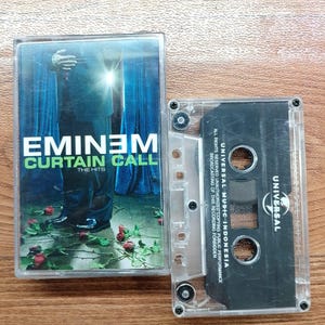Puede incluir: Un casete del álbum de Eminem "Curtain Call: The Hits". La portada del álbum presenta a un hombre con traje con el título del álbum en verde y blanco. El casete es transparente con una etiqueta negra.