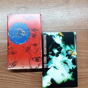Puede incluir: Dos cintas de cassette. Una tiene una portada roja con un círculo azul e ilustraciones de ojos, la palabra "wish" y el nombre de la banda "cure". La otra tiene una portada negra con el texto "THE CURE - DISINTEGRATION" y una imagen verde y blanca.