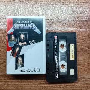Może przedstawiać: Vintage kaseta i etui zespołu Metallica. Etui jest biało-czerwone z logo zespołu i zdjęciami członków. Czarna kaseta jest oznaczona jako "BASF ferro super. AQUARIUS".