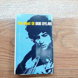 May include: A paperback book titled "The Best Of Bob Dylan" featuring a black and white image of Bob Dylan playing a guitar. The book has a light blue background with yellow text. The cover art is a portrait of the musician.