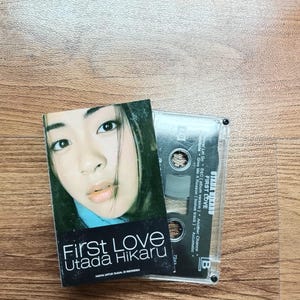 May include: A cassette tape and album cover for "First Love" by Utada Hikaru. The album cover features a close-up of a woman's face with the album title in white text. The cassette tape is clear.