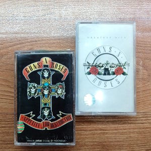 Puede incluir: Dos cintas de casete de Guns N' Roses. Una presenta la portada del álbum "Appetite for Destruction" sobre un fondo negro. La otra es una compilación de "Greatest Hits" con un fondo blanco y el logotipo de la banda.