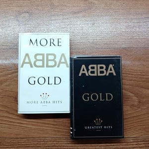Puede incluir: Dos cintas de casete de música de ABBA. Una es blanca con "MORE ABBA GOLD" impreso en dorado. La otra es negra con "ABBA GOLD GREATEST HITS" en dorado. Ambas tienen un diseño de corona dorada.