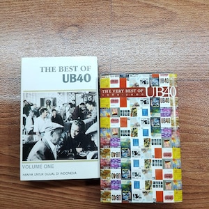 Puede incluir: Dos álbumes recopilatorios de música de UB40. Uno se titula "The Best Of UB40 Volume One" con una foto en blanco y negro. El otro es "The Very Best Of 1980-2000" con un collage colorido de imágenes. Sobre una superficie de madera.