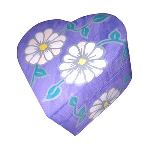 K&ouml;nnte beinhalten: Lilafarbener, herzf&ouml;rmiger Holzkasten mit Blumenmuster. Die Box ist mit wei&szlig;en G&auml;nsebl&uuml;mchen und gr&uuml;nen Bl&auml;ttern bemalt.