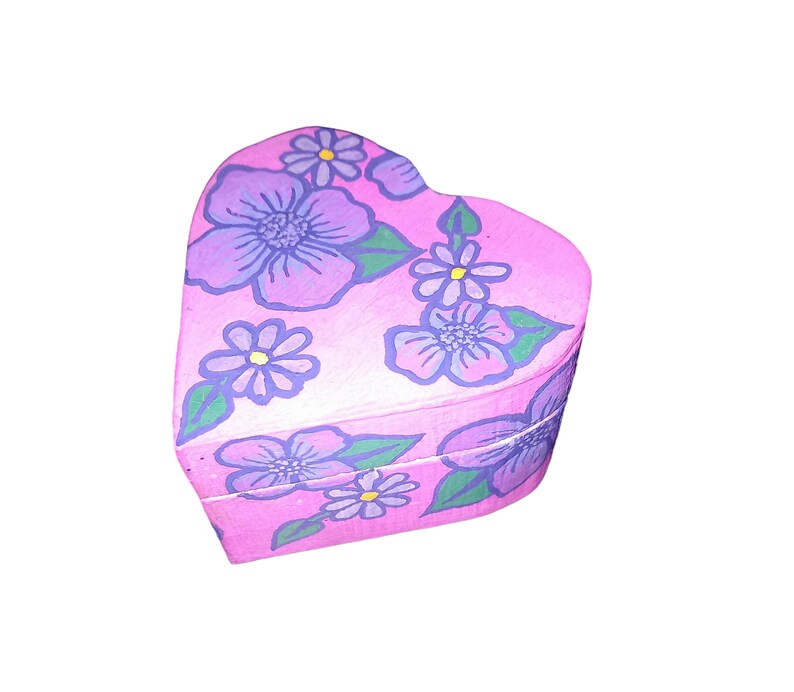 K&ouml;nnte beinhalten: Eine rosa herzf&ouml;rmige Schachtel mit einem Blumendesign. Die Blumen sind lila und blau mit gelben Zentren. Die Schachtel ist mit einem hellrosa Hintergrund bemalt.