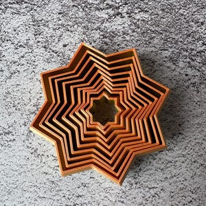 May include: A set of copper-colored star-shaped cookie cutters nested together. The cutters are arranged in a starburst pattern, with each cutter slightly smaller than the last. The cutters are made of a shiny metal and are sitting on a textured gray surface.