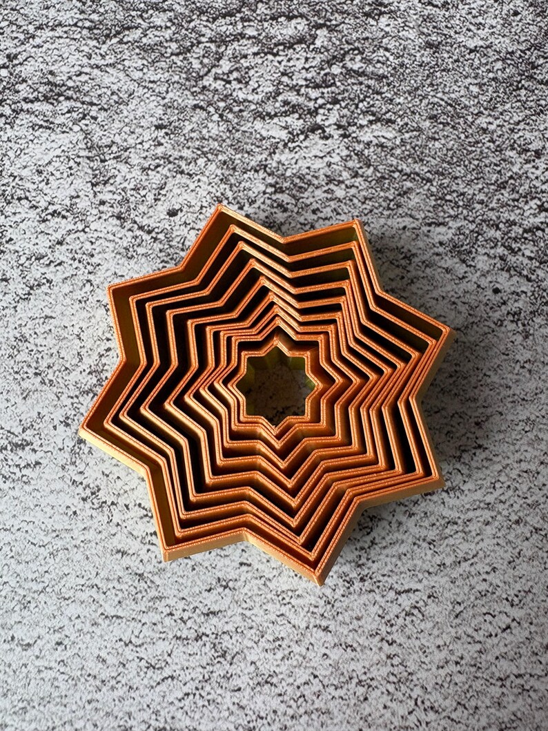 May include: A set of copper-colored star-shaped cookie cutters nested together. The cutters are arranged in a starburst pattern, with each cutter slightly smaller than the last. The cutters are made of a shiny metal and are sitting on a textured gray surface.