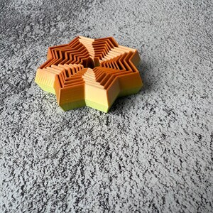 May include: A multi-layered, star-shaped object with a gradient of colors. The object transitions from light green to yellow, then to orange and brown. The design features a series of zig-zag edges, creating a unique, dimensional effect.