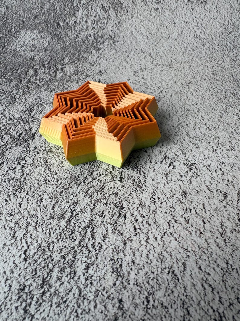 May include: A multi-layered, star-shaped object with a gradient of colors. The object transitions from light green to yellow, then to orange and brown. The design features a series of zig-zag edges, creating a unique, dimensional effect.