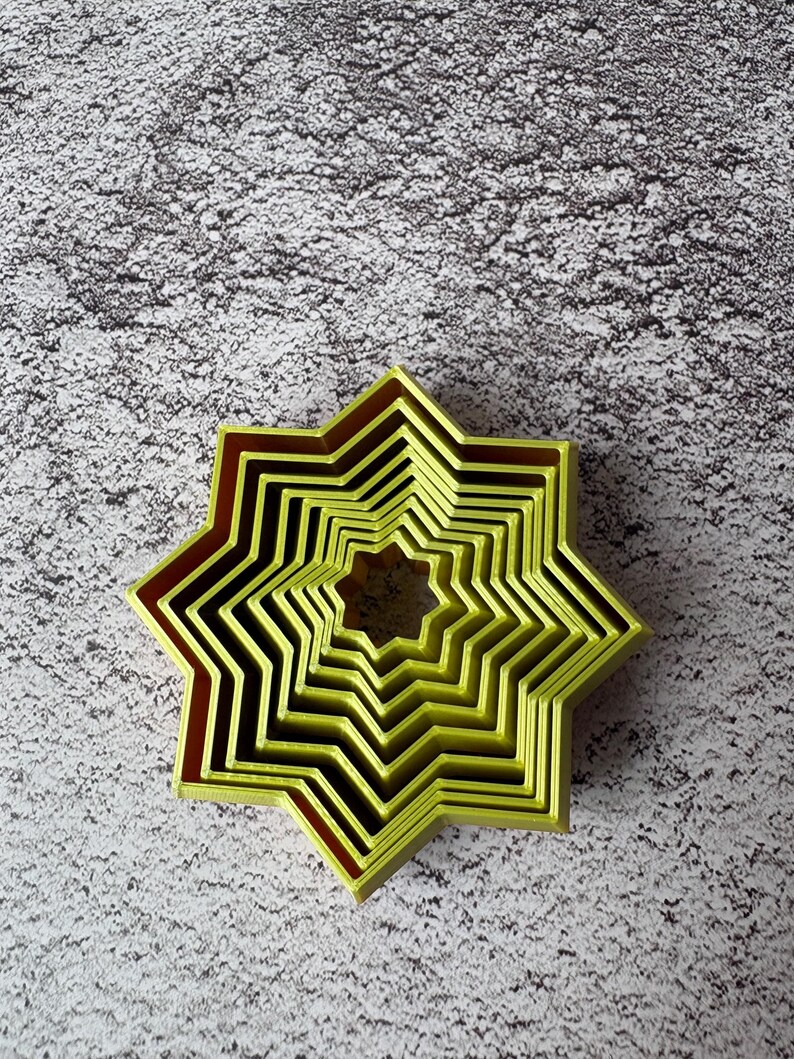 May include: A nested set of star-shaped cookie cutters in varying sizes. The cutters are a bright yellow-green color with a dark red-brown trim. The cutters are arranged in a nested pattern, with the smallest star in the center.