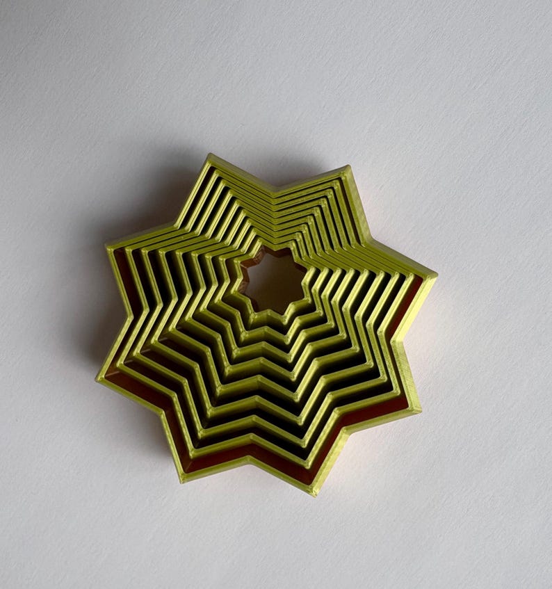 May include: A layered star-shaped object with a nested design. The object features a light green exterior with a dark red interior, creating a contrast. The star has multiple layers, giving it a three-dimensional appearance. The object is likely a decorative item.