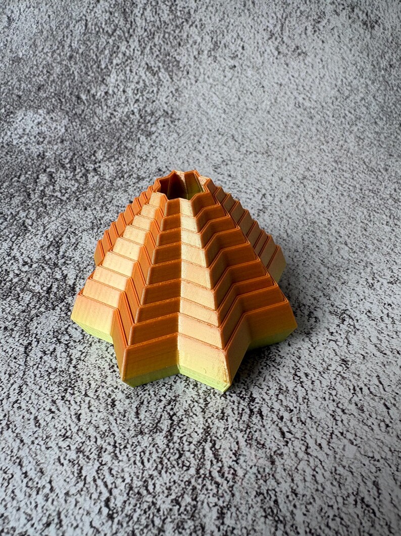 May include: A geometric, star-shaped decorative object with a stepped design. The object features a gradient of colors, transitioning from yellow-green at the base to orange at the top. The object is likely 3D-printed and has a unique, modern aesthetic.