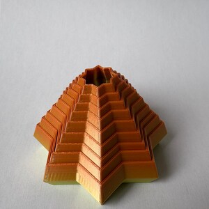 May include: An orange and yellow geometric pyramid-shaped object with a star-shaped base. The object has a layered design with a zig-zag pattern. The top has a circular opening. The object is likely 3D-printed.
