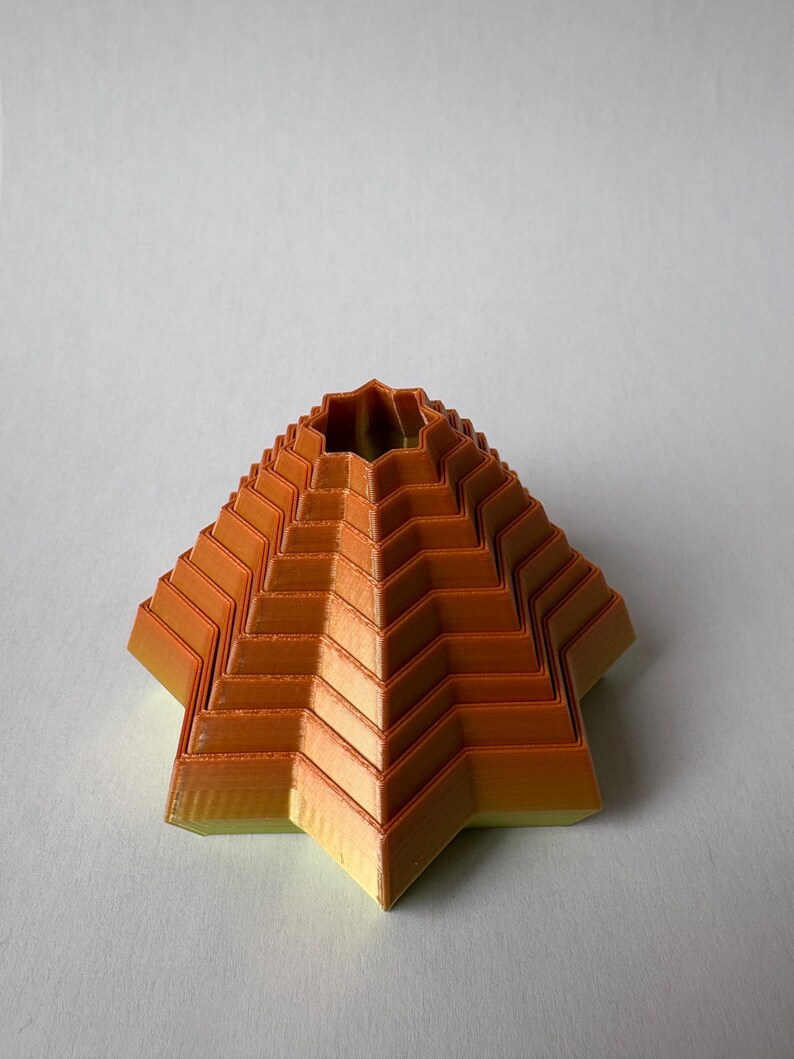 May include: An orange and yellow geometric pyramid-shaped object with a star-shaped base. The object has a layered design with a zig-zag pattern. The top has a circular opening. The object is likely 3D-printed.