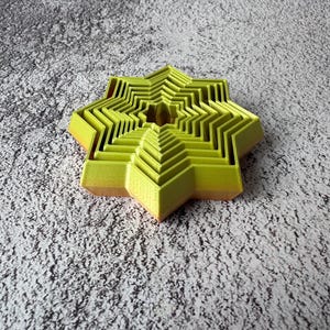 May include: A layered, star-shaped object in shades of yellow and orange. The object has a geometric design with multiple nested layers, creating a three-dimensional effect. The star is set against a textured, gray background.