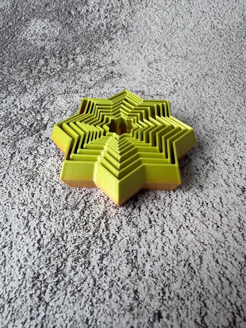 May include: A layered, star-shaped object in shades of yellow and orange. The object has a geometric design with multiple nested layers, creating a three-dimensional effect. The star is set against a textured, gray background.