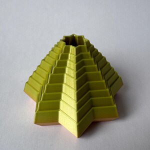 May include: A 3D-printed, geometric sculpture in a pyramid shape. The sculpture is a vibrant yellow-green color with a textured surface, featuring stepped layers. The base has a star-like shape. The object is set against a white background.