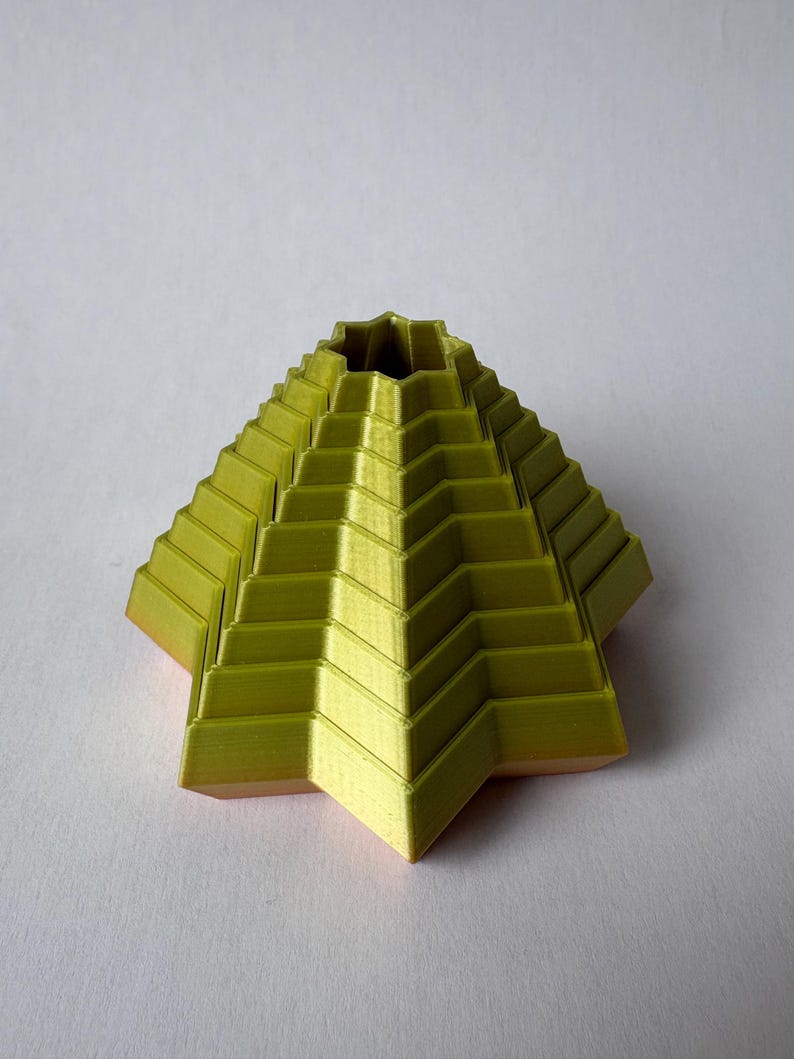 May include: A 3D-printed, geometric sculpture in a pyramid shape. The sculpture is a vibrant yellow-green color with a textured surface, featuring stepped layers. The base has a star-like shape. The object is set against a white background.