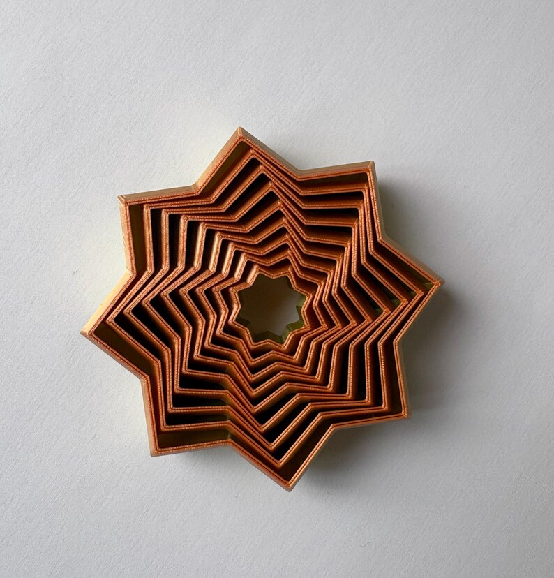 May include: A set of copper-colored star-shaped cookie cutters nested together. The cutters are arranged in a layered pattern, with each star slightly smaller than the last, creating a dimensional effect. The cutters are made of metal.