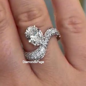 May include: A diamond ring with a large oval-cut center stone and a wave-like band encrusted with smaller diamonds. The ring is set in a silver metal and is displayed on a hand. The text "DiamondsPage" is visible.