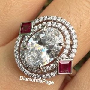 May include: A silver ring featuring a large, oval-cut diamond surrounded by a double halo of smaller diamonds. Two square, red gemstones flank the central stone. The ring is set against a blurred background. The text "DiamondsPage" is visible.