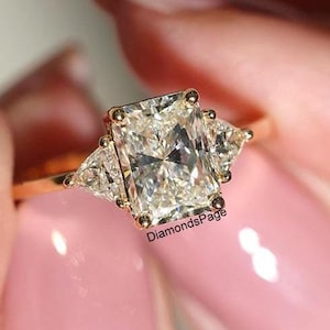 May include: A gold ring featuring a large, rectangular, clear diamond in the centre and two smaller, triangular, clear diamonds on either side. The ring is on a finger.
