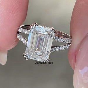 May include: A silver-toned ring featuring a large emerald-cut centre stone and a split shank band encrusted with small round pavé-set stones. The metal band has a polished finish and the text DiamondsPage is visible at the base of the setting.