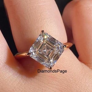 Peut inclure: Une bague avec une pierre précieuse transparente à facettes de taille carrée montée sur un anneau en or à quatre griffes. La pierre présente un motif distinctif de taille à degrés avec un point central. Le texte DiamondsPage est inscrit.
